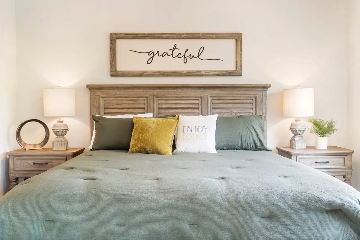 Another king bedroom showcasing a bed with decorative pillows, a sign reading 'grateful' above the headboard, and matching nightstands with softly glowing lamps. The neutral color palette and soft lighting evoke a sense of calm and reflection.