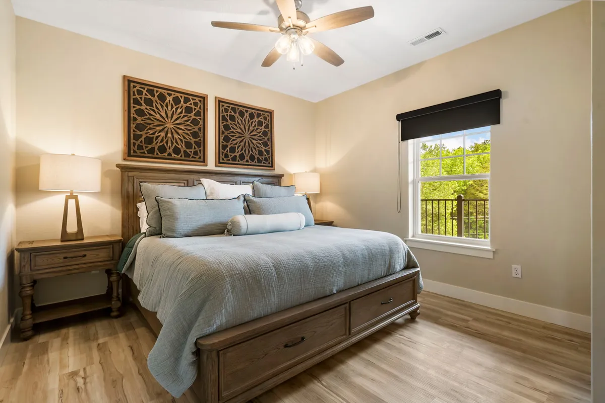 A serene king bedroom featuring a bed with decorative pillows and a carved wooden headboard, illuminated by natural light streaming through a nearby window. Bedside lamps and warm wood accents create an inviting, peaceful atmosphere ideal for privacy and relaxation.