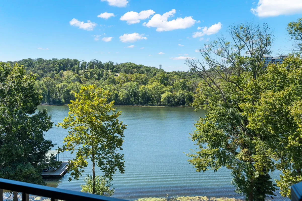 The image presents a serene panorama of Lake Taneycomo, with its glassy, calm water stretching toward lush green trees and rolling distant hills. The inviting landscape hints at opportunities for boating, fishing, and peaceful nature walks, while the expansive view encourages guests to unwind and soak in the tranquility of the lakeside setting.