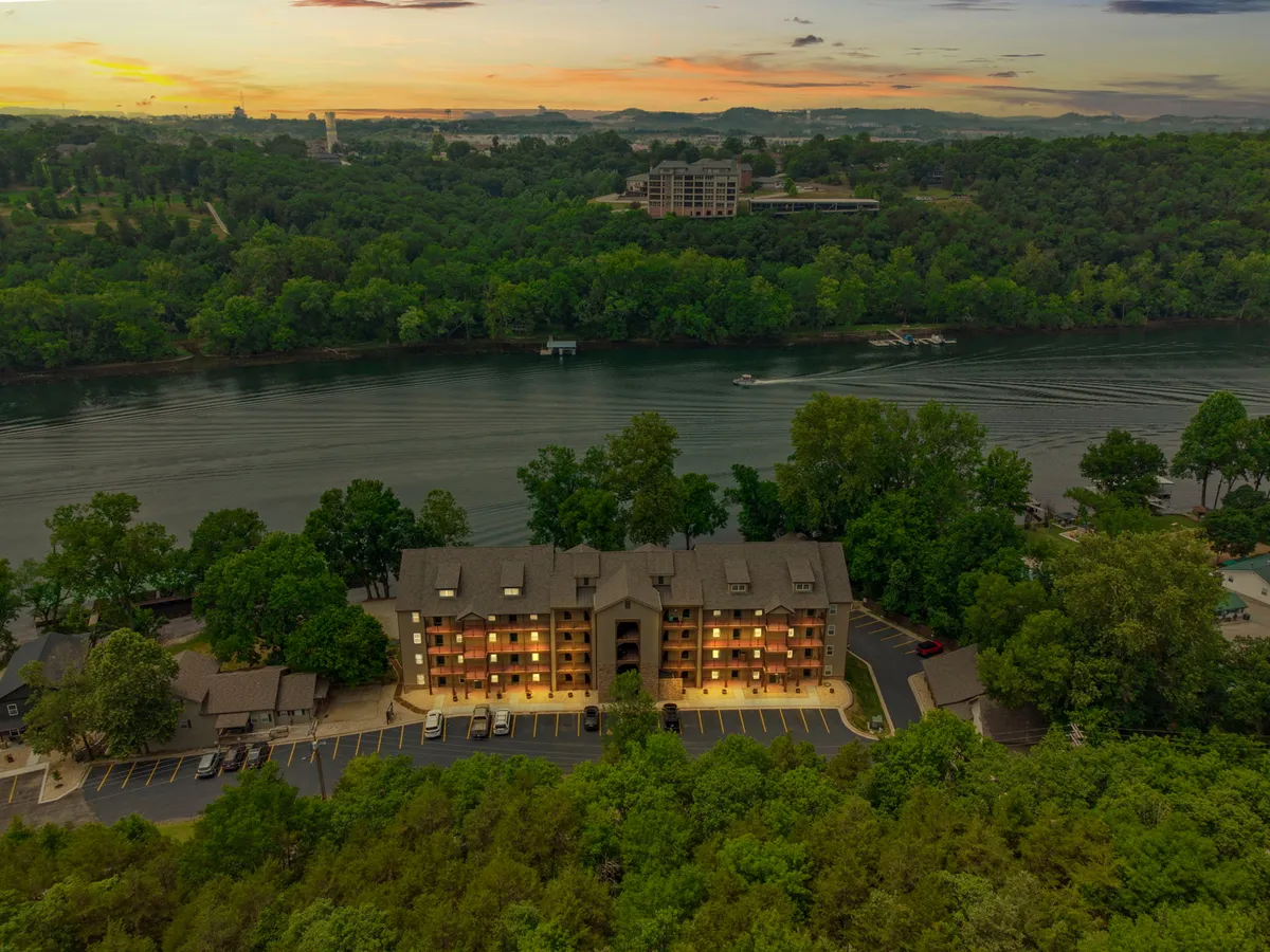 This exterior shot captures the multi-story condo building situated right beside the river, framed by lush greenery and set beneath a glowing sunset sky. The proximity to the water and the surrounding landscape is clearly visible, conveying a tranquil and attractive setting.