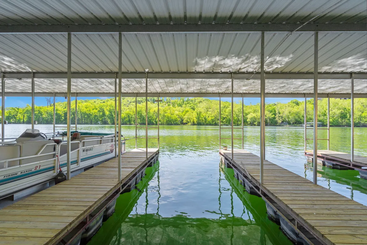 This image captures pontoon boats moored at wooden docks with lush green trees reflected in the calm waters of the lake. The setting suggests opportunities for boating, fishing, and picnicking directly by the lake, with the tranquil water and surrounding greenery evoking a peaceful, family-friendly atmosphere.
