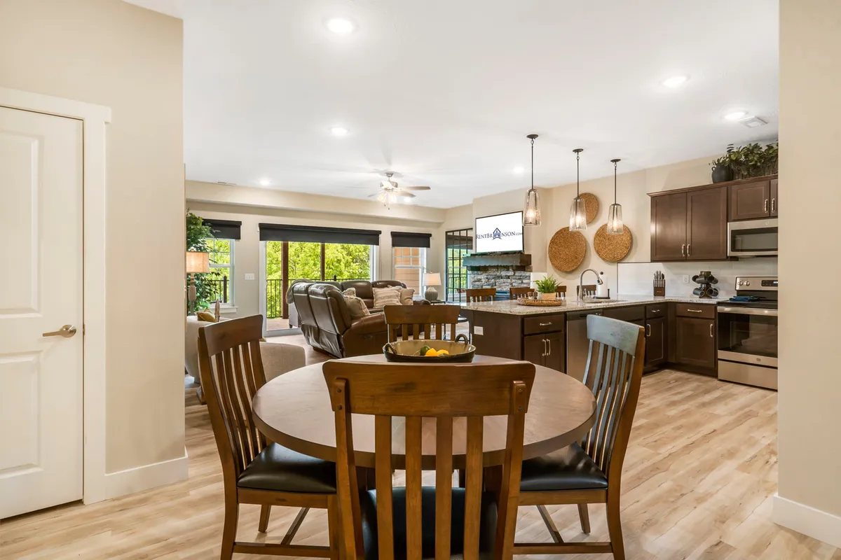 A dining table with a vibrant fruit centerpiece sits beside the kitchen area, with cozy living room seating and windows allowing natural light to fill the space. The scene exudes warmth, suggesting inviting family meals and celebrations.